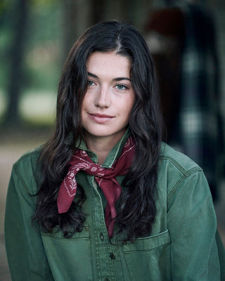 A woman with long dark wavy hair wearing an olive green button-up shirt and a maroon paisley neck scarf, photographed outdoors against a softly blurred background.