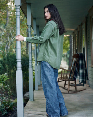 A woman stands on a wooden porch wearing an olive green long-sleeve button-up shirt and wide-leg blue jeans, holding onto a gray post.