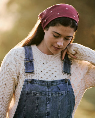 A woman wearing a cream cable-knit sweater, blue denim overalls, and a burgundy bandana with small white star details, set against a warm, blurred outdoor background.