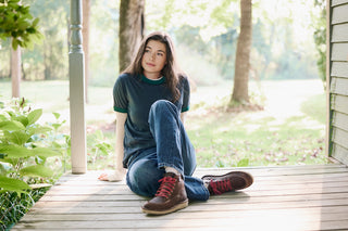 A young woman sits on a wooden porch deck wearing a dark green t-shirt, blue jeans, and brown lace-up boots with red laces, set against a soft-focus background of trees and grass.