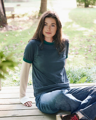 A woman sits on a wooden deck in an outdoor park setting, wearing a dark teal short-sleeve t-shirt with green trim, blue jeans, and red sneakers.