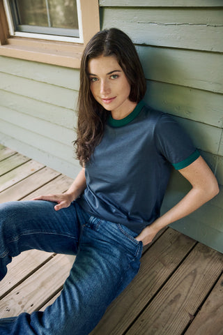 A woman sitting on a wooden deck against a light green house wall, wearing a slate blue ringer t-shirt with a dark green crew neck and sleeve trim, paired with blue jeans.