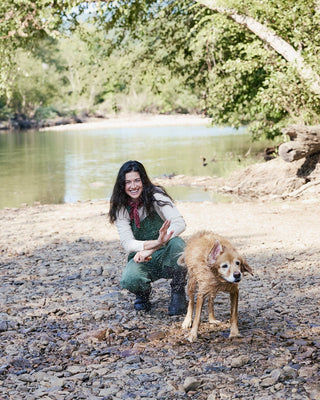 A woman crouches on a rocky riverbank next to a dog, with a calm river and green trees in the background.