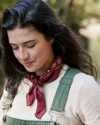 A woman outdoors wearing green denim overalls over a cream knit top, with a dark red paisley neck scarf and dark brown hair.