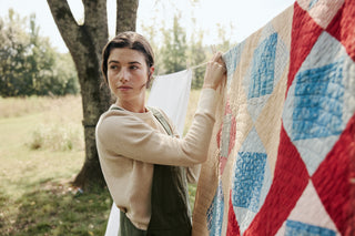 A woman outdoors hanging a quilted patchwork blanket with red, blue, beige, and white geometric blocks next to a tree.