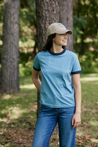 A woman wearing a light blue ringer t-shirt with dark navy trim, blue jeans, and a beige baseball cap, standing in a wooded outdoor setting.