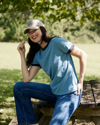 A woman sitting on a wooden picnic table outdoors, wearing a light blue ringer t-shirt with dark navy trim, blue jeans, and a beige baseball cap.