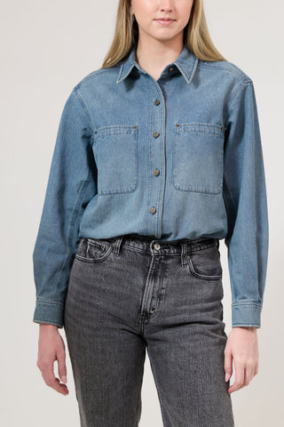 Person wearing a blue denim shirt and dark jeans on a plain background