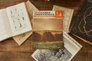 Vintage magazine titled 'The Ozarks Mountaineer' on a wooden surface with other books and papers.