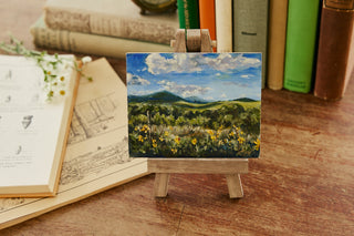 Miniature landscape painting on a small easel with books and an open notebook in the background.