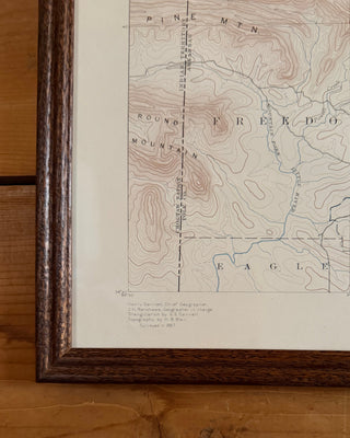 A framed vintage topographic map print resting on a rustic wooden table. The map features fine brown contour lines, blue river details, and a dark brown wooden frame.