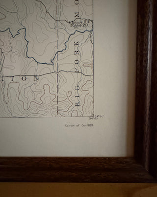 A close-up of a framed vintage map print showing topographic contour lines, place names, and a small caption reading 'Edition of Oct 1899.' The print is mounted on off-white paper inside a dark brown wooden frame.