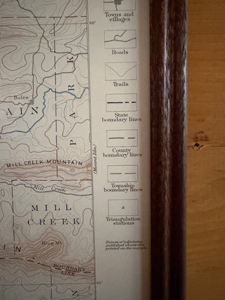 A close-up of a vintage topographical map showing Mill Creek and Mill Creek Mountain with contour lines, place names, and legend symbols, positioned next to a dark wooden frame on a wood surface.