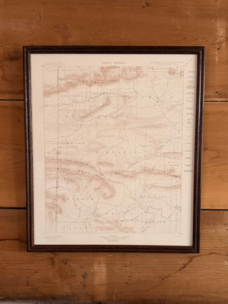A framed vintage topographical map print with fine pink and blue line details on aged cream paper, enclosed in a dark brown wooden frame and mounted on a rustic wooden plank wall.
