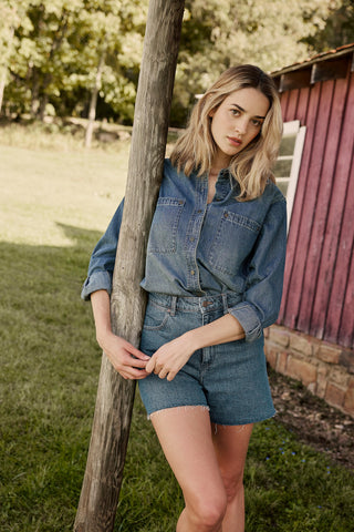 Woman in denim outfit leaning against a wooden post outdoors.