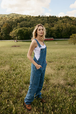 Woman wearing blue overalls standing in a grassy field with trees and a blue sky in the background