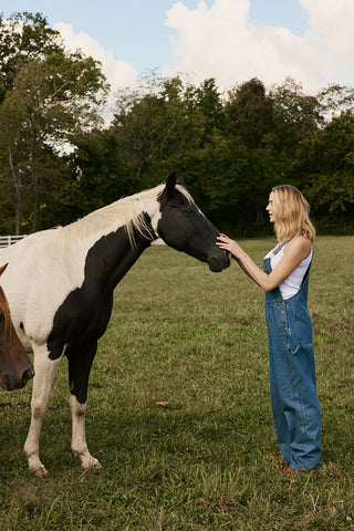 A woman in blue denim overalls stands in a grassy field, gently touching the face of a black-and-white paint horse. Another horse is partially visible on the left, with trees and a cloudy sky in the background.