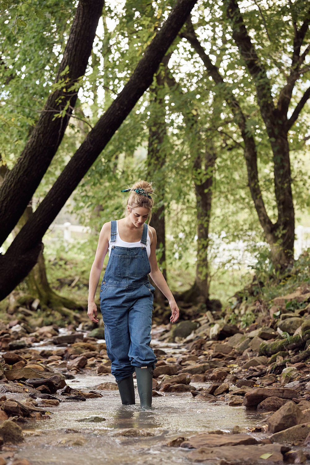 Person walking through a stream in a forest wearing blue overalls and rain boots.