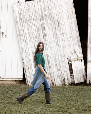 A woman walks across a grassy field in front of a large, weathered white barn door. She is wearing a dark green short-sleeve t-shirt, a pair of blue denim overalls over blue jeans, and dark brown rubber boots.