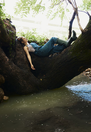 Person lying on a large tree trunk by a body of water with greenery in the background